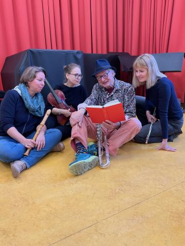 Wollie Kaiser und Ensemble sitzen auf dem Boden mit Buch und Musikinstrumenten